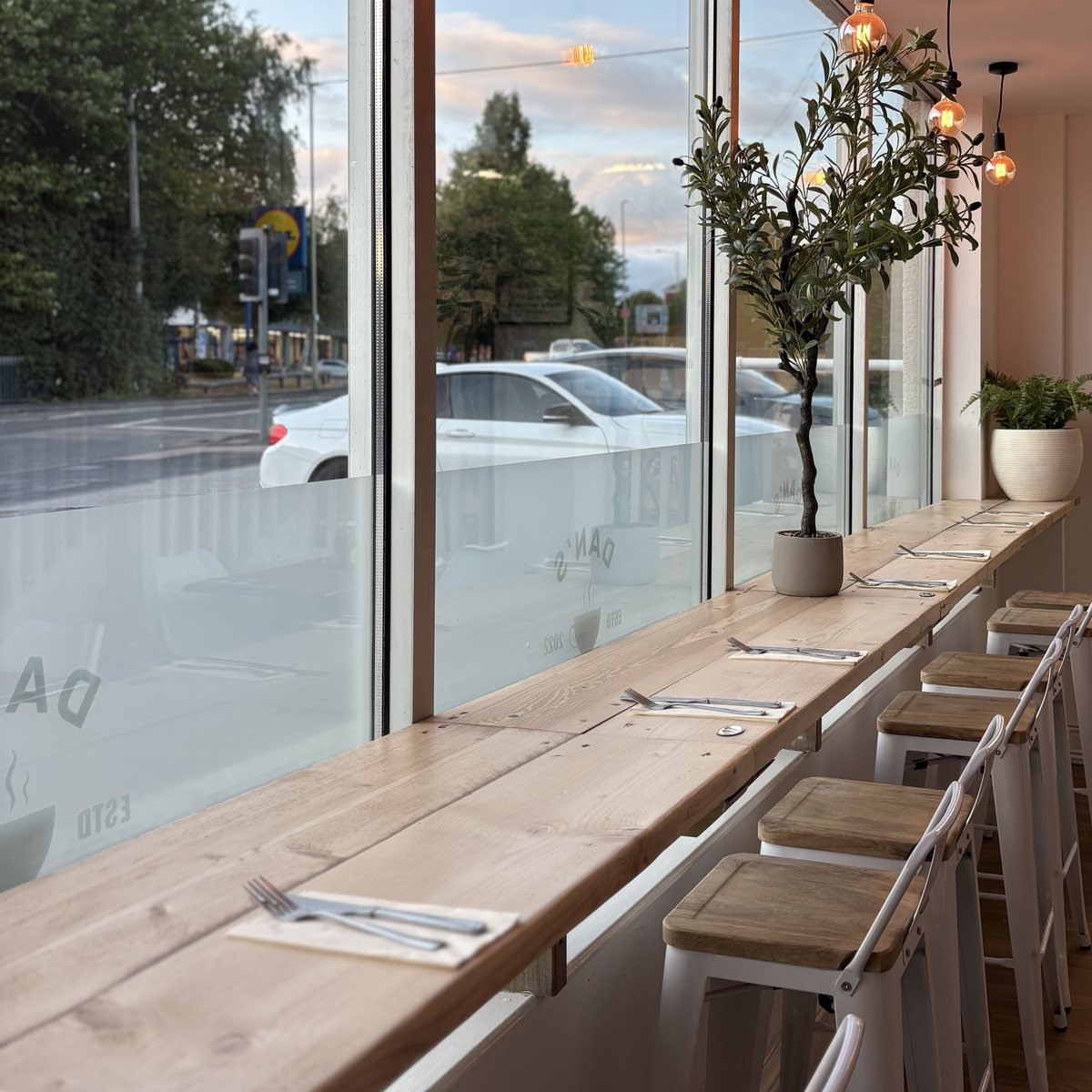 The window bar at Dan's Cafe at dusk, set with cutlery beneath warm Edison bulbs, with an olive tree and the etched Dan's Cafe logo on the glass